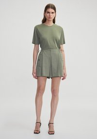 Camiseta de algodón de color verde oliva con mangas cortas combinada con pantalones cortos plisados de color verde oliva que cuentan con bolsillos laterales y detalles decorativos.