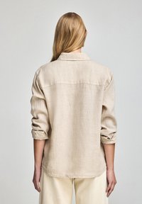Camisa de lino beige con cuello, mangas remangadas y dobladillo recto. Presenta una superficie texturizada y un diseño sencillo y minimalista.