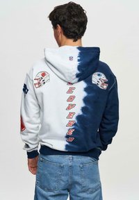 Sweat-shirt à capuche présentant un mélange de teinture blanche et bleu marine, avec des logos NFL et le texte "COVERED" au dos. En coton.