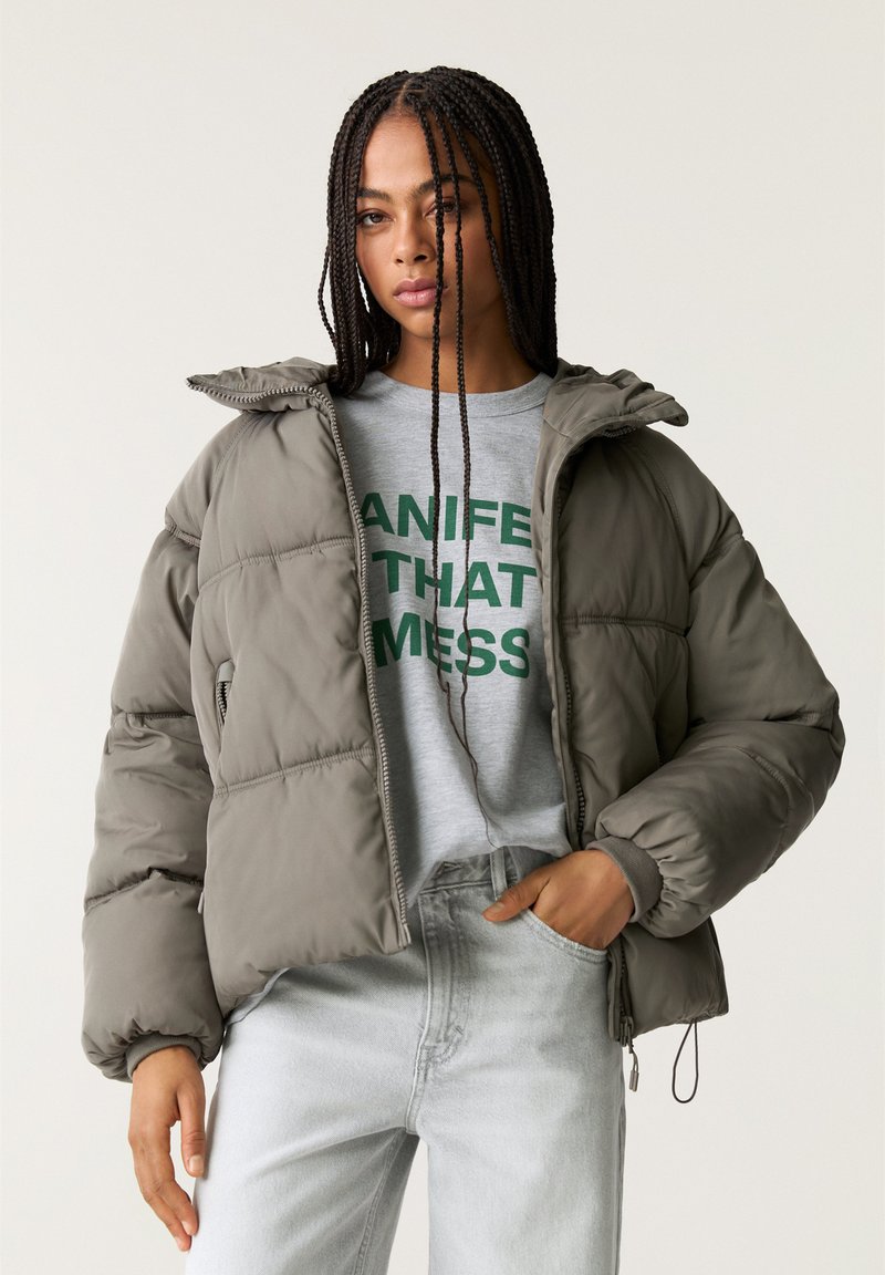 Giacca a vento in verde oliva spento, con chiusura a zip, cappuccio e design trapuntato. Indossata sopra una felpa grigia con testo verde.