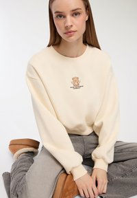 Felpa cremosa con scollo rotondo e polsini aderenti, con una grafica di un orso con ali e testo. Abbinata a jeans grigi a gamba larga e scarpe color sabbia.