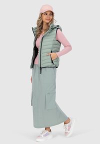 Gilet verde chiaro con spalle imbottite e tasche, abbinato a una maglietta rosa a maniche lunghe e un berretto rosa coordinato. Gonna lunga beige e sneakers bianche con dettagli.