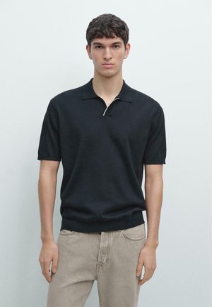 Jeune homme aux cheveux courts et foncés portant une chemise noire à manches courtes avec col et un pantalon beige, debout devant un fond clair uni.