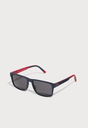 Óculos de sol de armação quadrada feitos de plástico azul-marinho com lentes cinzentas, apresentando detalhes em vermelho nas hastes e uma superfície texturizada.