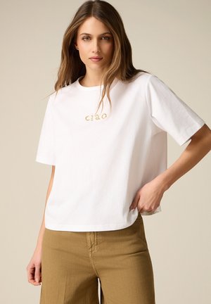 Femme aux longs cheveux portant un t-shirt blanc avec le texte « ciao » et un pantalon marron, posant avec une main dans sa poche.