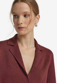 Burgunderfarbene strukturierte Strickjacke mit Reverskragen und offenem Ausschnitt. Das Model trägt silberne Creolen. Schlichter Hintergrund.