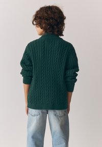 Suéter verde de punto grueso con cuello alto y mangas remangadas, combinado con vaqueros de mezclilla azul claro. La textura es gruesa y cálida.