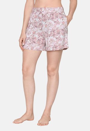 Vrouw draagt pastelroze korte broek met bloemenprint, elastische taille en trekkoord, staat op blote voeten met één hand in de zak.