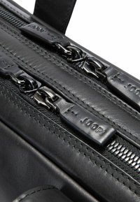 Schwarze Ledertasche mit metallischen Reißverschlüssen und Zuglaschen. Auffällige Nähte und Texturdetails heben die Qualität des Materials hervor.