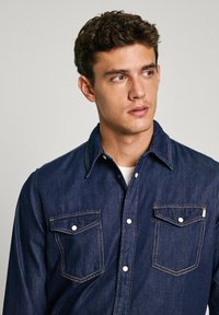 Donker denimshirt met twee borstzakken, contrasterende stiksel en een klassieke kraag. De stof heeft een soepele textuur en gestructureerde pasvorm.