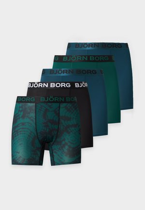 Homme torse nu, debout pieds nus, portant un boxer sombre à motifs avec le logo Björn Borg sur la ceinture, sur un fond clair uni.