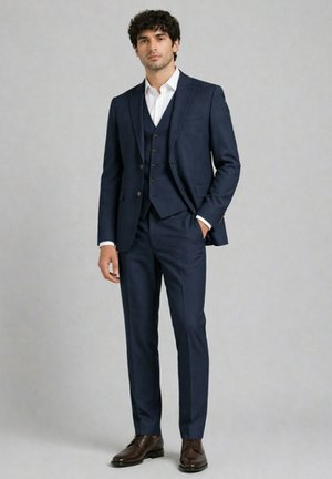 Homme debout portant un costume trois pièces bleu marine, une chemise blanche et des chaussures marron sur un fond gris clair uni.