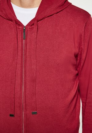 Rote Zip-Hoodie aus strukturiertem Strick, mit Kapuze, Kordelzug und schwarzen Hardware-Akzenten. Langärmliges Design, getragen über einem weißen Shirt.