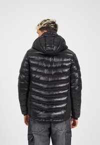 Črna puhovka z sijočim zaključkom, horizontalno prešita zasnova, pritrjena kapuca in elastični manšeti. Material se zdi lahek in sijoč.