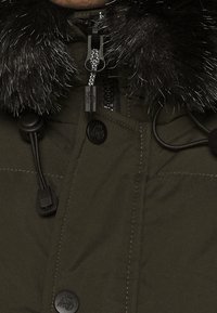 Parka verde con capucha forrada de piel negra, cierres de cremallera y broches a presión, cordones ajustables y tejido texturizado.