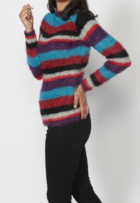 Pull en tricot à rayures multicolores avec une texture duveteuse ; les couleurs incluent bleu, rouge, violet, noir et gris. Col rond et manches longues.