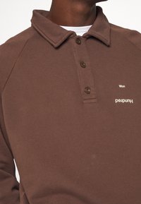 Polo shirt in cotone marrone con colletto, tre bottoni e stampa "Hundred" sul petto. Tessuto texturizzato e maniche raglan.