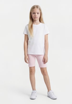 Mädchen mit langen blonden Haaren, das steht, trägt ein einfaches weißes T-Shirt, helle rosa Shorts und weiße Turnschuhe vor einem weißen Hintergrund.