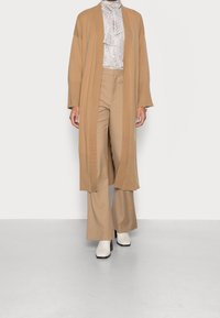 Cardigan lungo beige con maniche larghe sopra una maglietta a fantasia bianca e nera, abbinato a pantaloni beige a gamba larga e stivali bianchi con tacco quadrato.