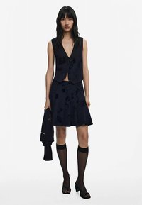 Tumesinine vestikujuline top sügava dekolteega, kombineeritud lillemustriga lehviva seelikuga. Modell kannab läbipaistvaid musti põlvikuid ja avatud varvastega kontsakingi.