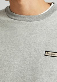 Felpa grigia con collo a giro, caratterizzata da un'etichetta tessuta con "Pepe Jeans" in corsivo, posizionata su una superficie di tessuto liscia e morbida.