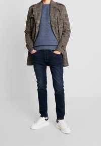 Abrigo a cuadros marrón y negro sobre un suéter azul texturizado, combinado con jeans oscuros y zapatillas blancas con detalles en azul marino.