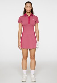 Polo-klänning i rosa tyg med korta ärmar, krage och sidoflikar. Styla med en vit logotyp, tillsammans med en handske och sportiga skor.