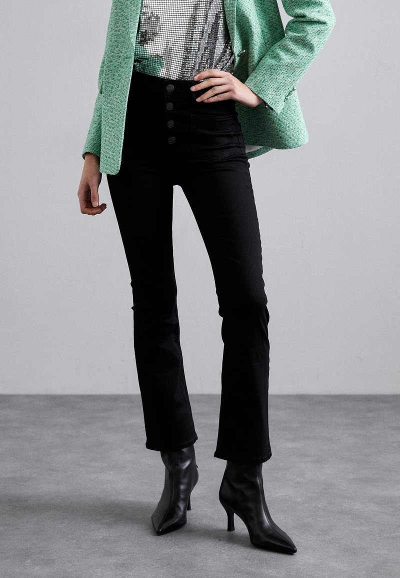 Jean flare noir avec taille haute à cinq boutons, associé à un blazer texturé vert et des escarpins noirs pointus, offrant une coupe soignée.