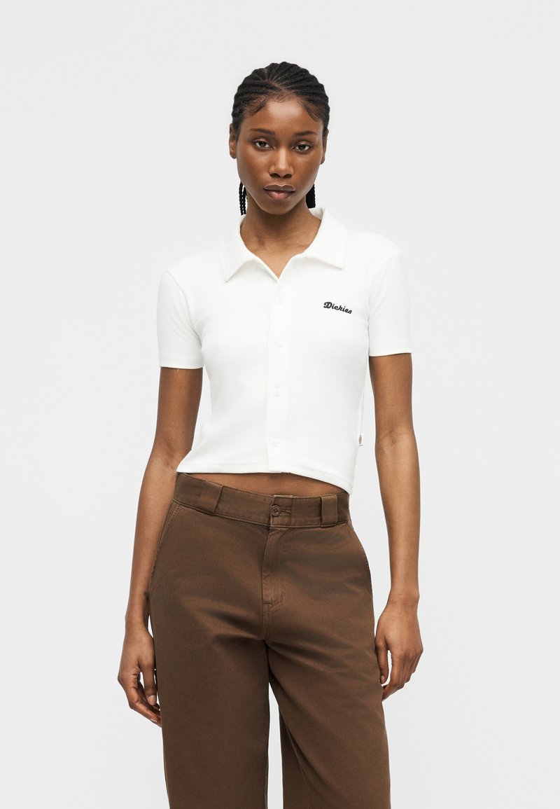 Giovane donna con capelli intrecciati che indossa un crop top bianco a maniche corte con colletto e pantaloni marroni a vita alta, in piedi contro uno sfondo semplice.