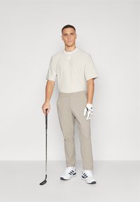 Cămașă de golf bej deschis cu mâneci scurte și guler alb, purtată cu pantaloni bej deschis. Mânușa de golf albă pe mâna stângă, ținând un club de golf.