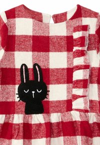 Punainen ja valkoinen ruudullinen kangasmekko, jossa on röyhelöitä, ja etupuolella musta brodeerattu pupu suljetuilla silmillä.