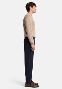 Beiges Polohemd mit langen Ärmeln, kombiniert mit maßgeschneiderten marineblauen Hosen; mit seitlicher Reißverschlusstasche und braunen Loafers. Schlichtes, modernes Design.