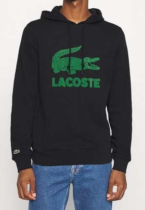 Sweat à capuche noir avec logo crocodile Lacoste vert et texte centré sur la poitrine, porté par une personne en jean bleu sur un fond uni.