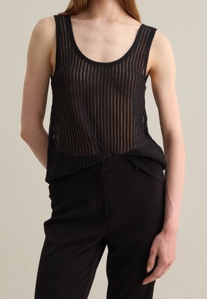 Femme portant un débardeur en maille noire sans manches et un pantalon noir, debout avec une main dans le dos sur un fond uni.