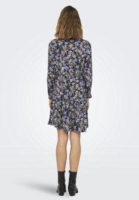 Vestito floreale con sfondo navy, caratterizzato da fiori viola e blu, maniche lunghe e orlo a strati. Indossato con stivali neri alla caviglia.