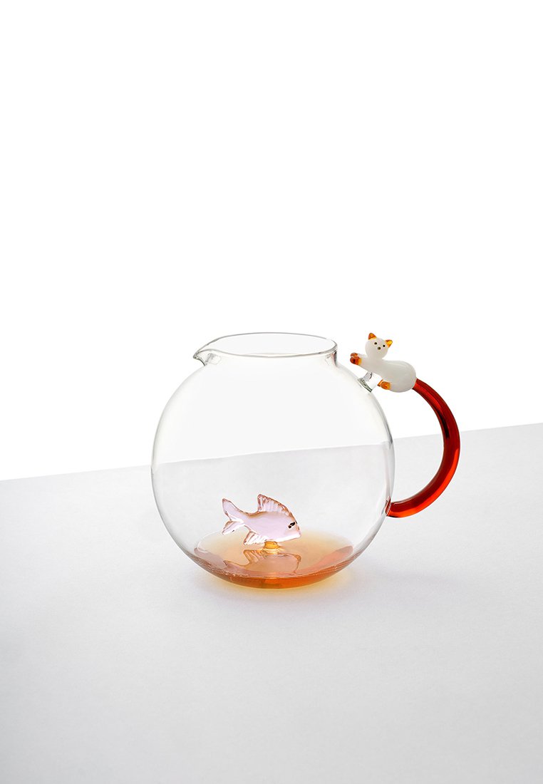 Przezroczysta szklana czajnik z czerwoną rączką, w którym znajduje się mały biały kot, umieszczony na dnie czajnika różowy szklany rybka, na białej powierzchni.