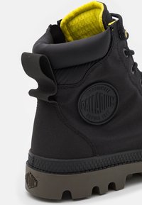 Bota de paladio negra con suela gruesa marrón, cuello acolchado en el tobillo, forro interior amarillo y logotipo de la marca en el lateral.