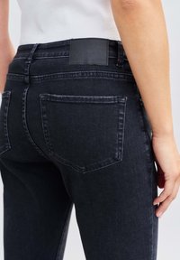 Gros plan d'une personne portant un jean en denim noir avec une poche arrière et un patch en cuir noir sur la ceinture, associé à un haut blanc.