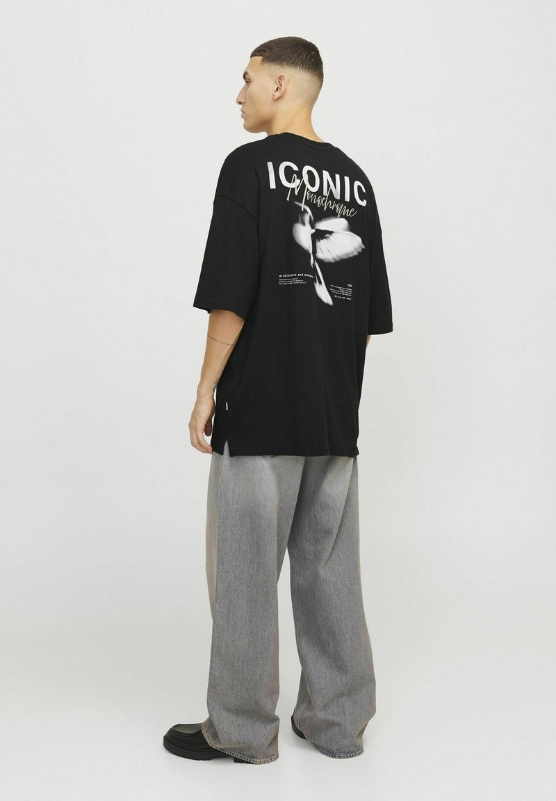 Czarna, oversize koszulka z graficznym nadrukiem, tekstem "IKONICZNY Monochrom" oraz białym motylem na plecach. Luźne dżinsy.