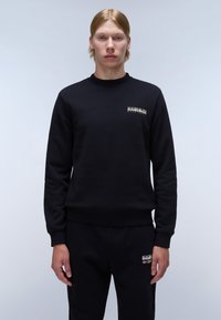 Sudadera negra con puños y cuello acanalados. Presenta un pequeño logo blanco en el lado izquierdo del pecho. Modelo vistiendo pantalones negros a juego.