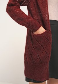 Burgunderfärgad stickad cardigan med öppen front, långa ärmar och diamantmönster. Har en ficka på vänster sida. Mjuk känsla.