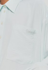 Helles blaues, strukturiertes Hemd mit Kragen, das eine Brusttasche und ein gesticktes "Calvin Klein"-Logo in passendem Faden aufweist.