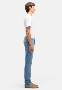 Jeune homme debout de profil portant un t-shirt blanc, un jean bleu et des bottines en suède beige sur un fond blanc uni.