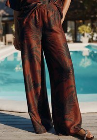 Pantalons larges en tissu marron rouille avec un motif de feuille de palmier noir. Taille élastique, texture lisse et design fluide.