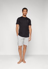 Zwarte t-shirt met korte mouwen en een klein logo op de borst, gecombineerd met lichtgrijze sportshorts. Model staat op blote voeten op een neutrale achtergrond.