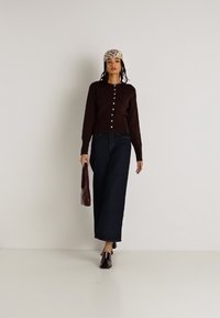 Cardigan castanho com botões, jeans largos escuros, sapatos bordo e um lenço de padrão. Modelo a segurar uma bolsa vermelha. Fundo neutro.