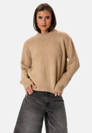 Femme aux cheveux bruns mi-longs portant un pull en maille beige et un jean ample gris foncé, debout les mains dans les poches.