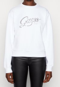 Felpa bianca con scollatura arrotondata, caratterizzata da un logo "Guess" in argento luccicante e il dettaglio "1981", abbinata a pantaloni in pelle nera.