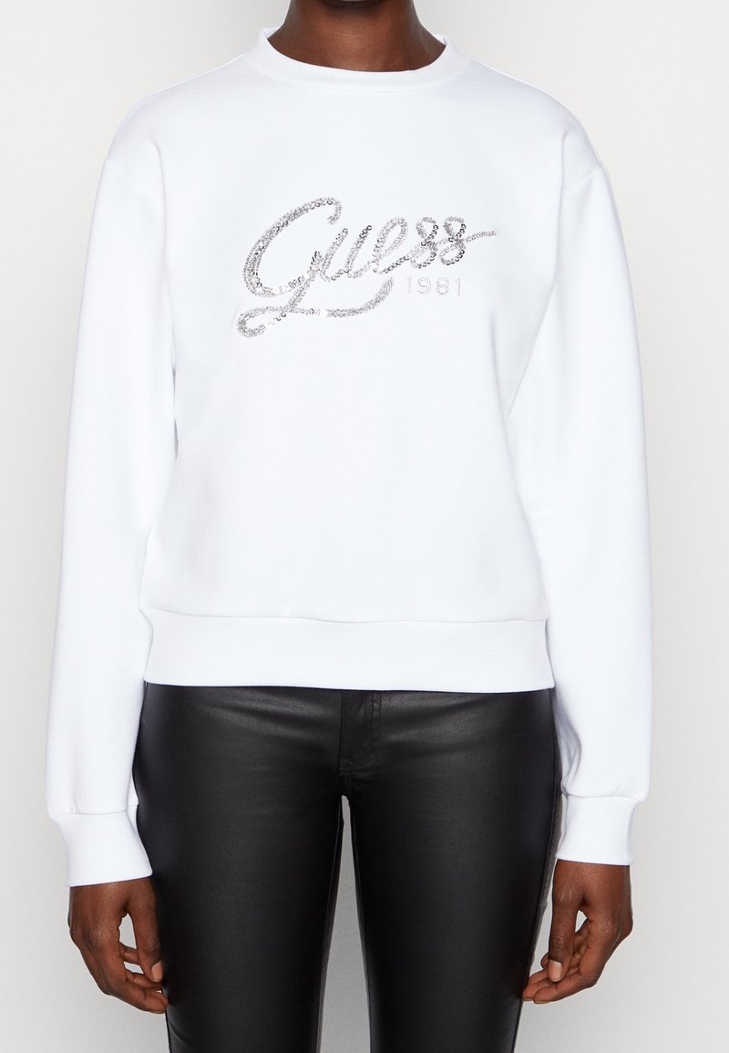 Felpa bianca con scollatura arrotondata, caratterizzata da un logo "Guess" in argento luccicante e il dettaglio "1981", abbinata a pantaloni in pelle nera.