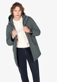 Joven con un abrigo de invierno gris con capucha sobre una sudadera crema y pantalones oscuros, sosteniendo los bordes del abrigo, mirando al frente contra un fondo blanco.